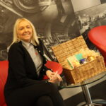 angela owen is sitting beside a weaved basket with fortnum and mason items inside it for the hamper raffle