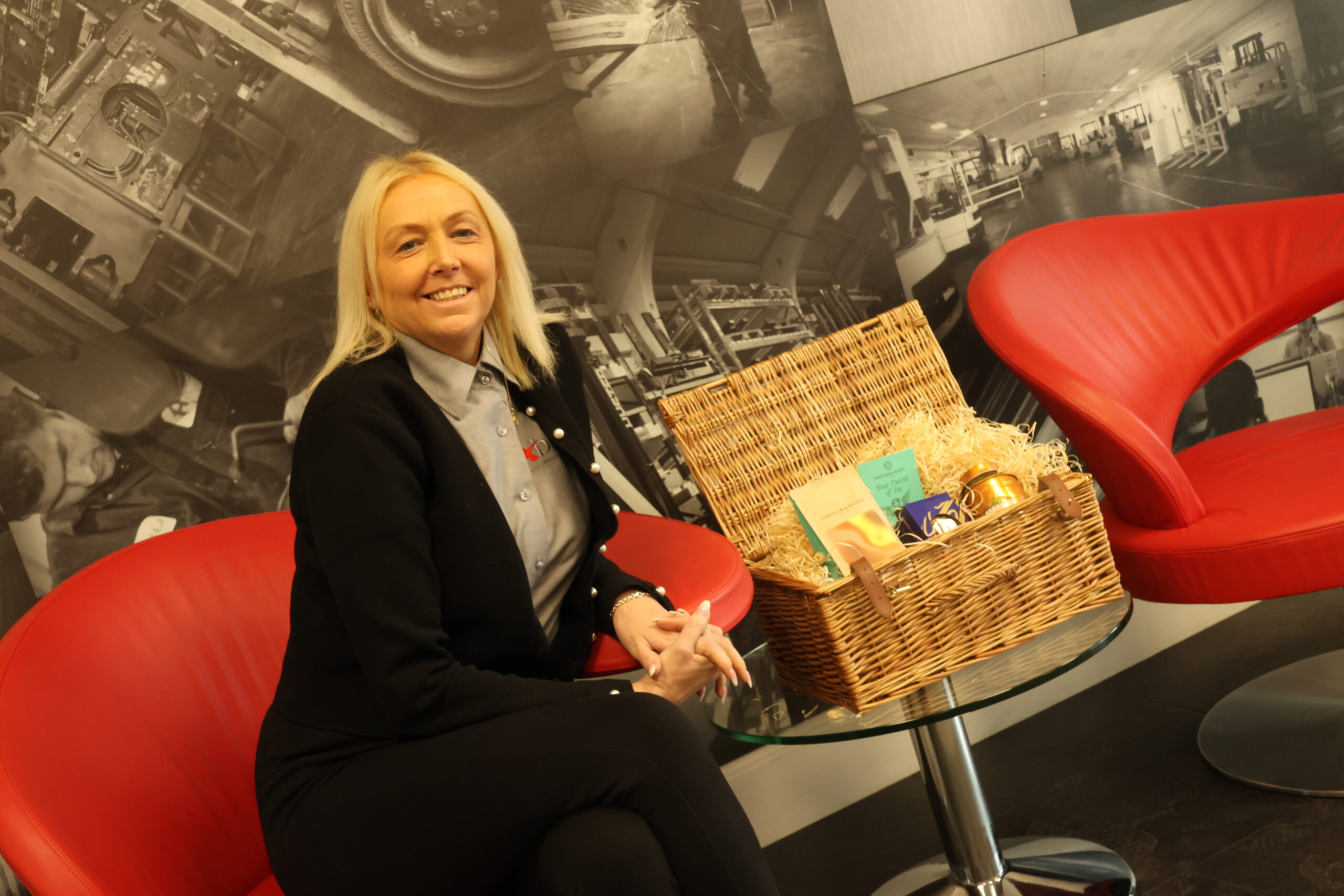 angela owen is sitting beside a weaved basket with fortnum and mason items inside it for the hamper raffle