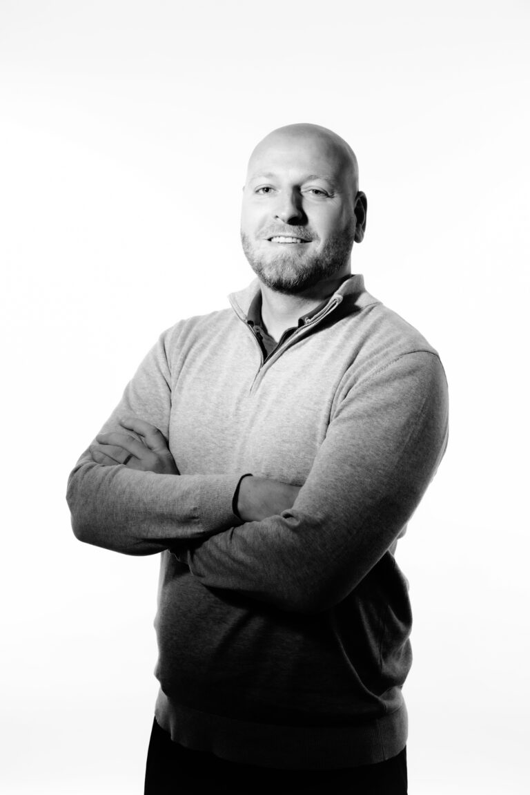 mark anderton is wearing a grey jumper with his arms folded whilst grinning at the camera.