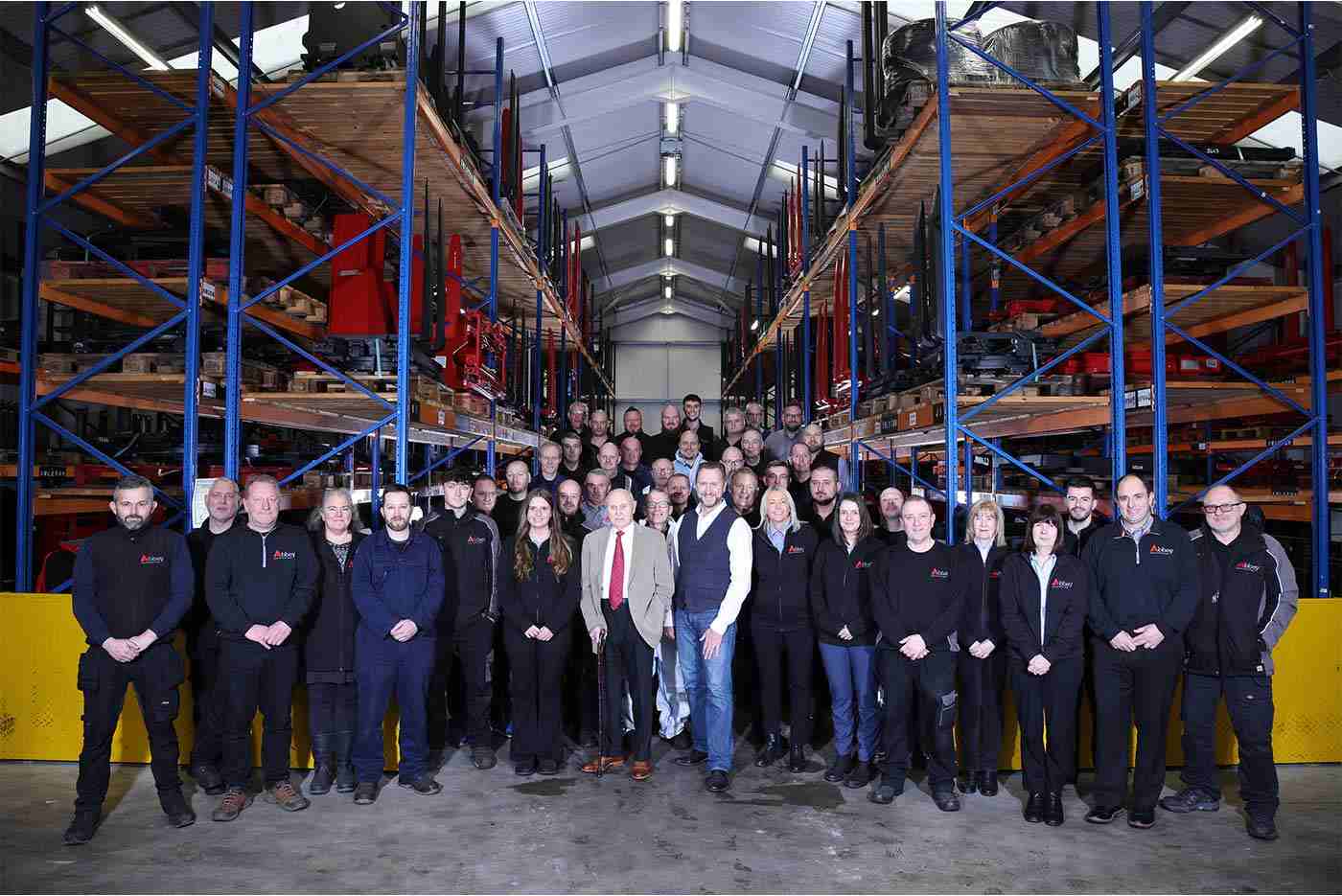 the entire abbey team wearing the black abbey jumpers are in a large warehouse surrounded by racking shelves