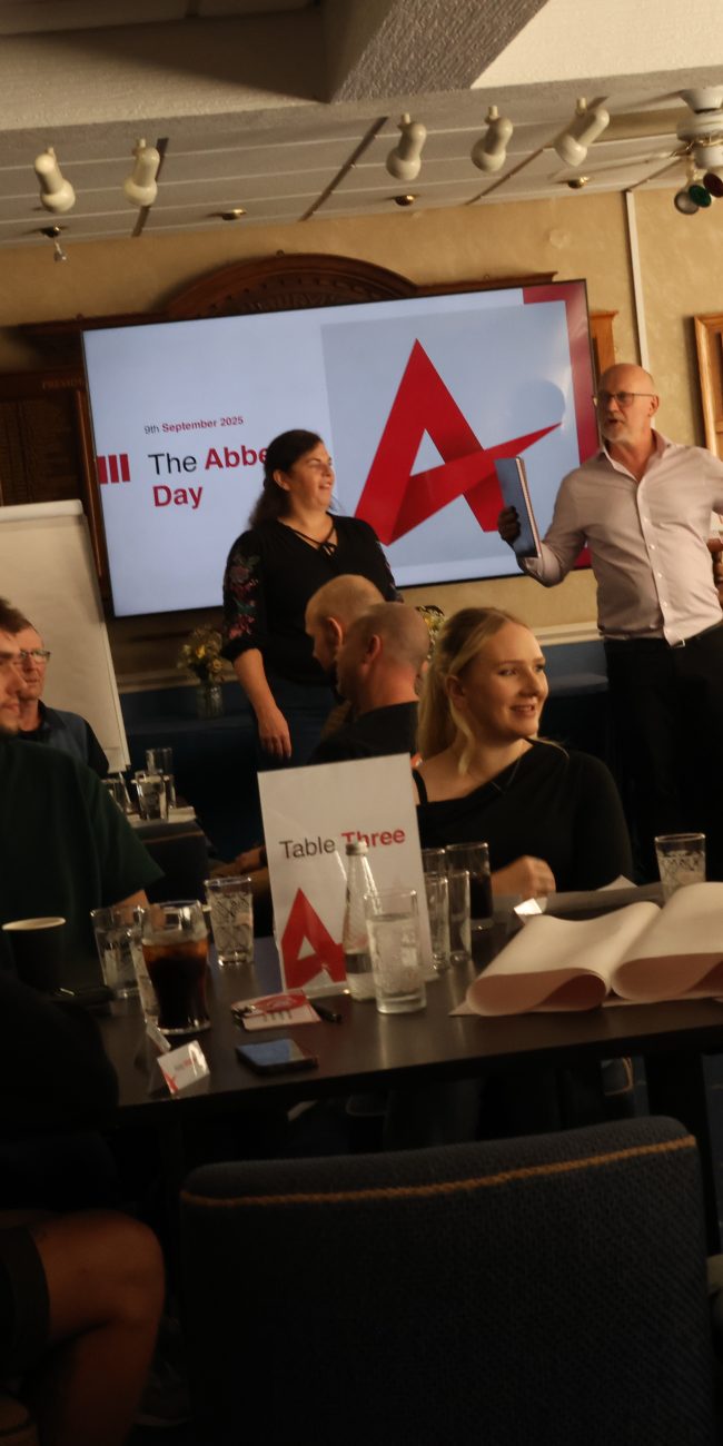 A collection of square tables with people sitting at them at smiling with a large TV behind them with 'The Abbey Day 2025' emblazoned onto it