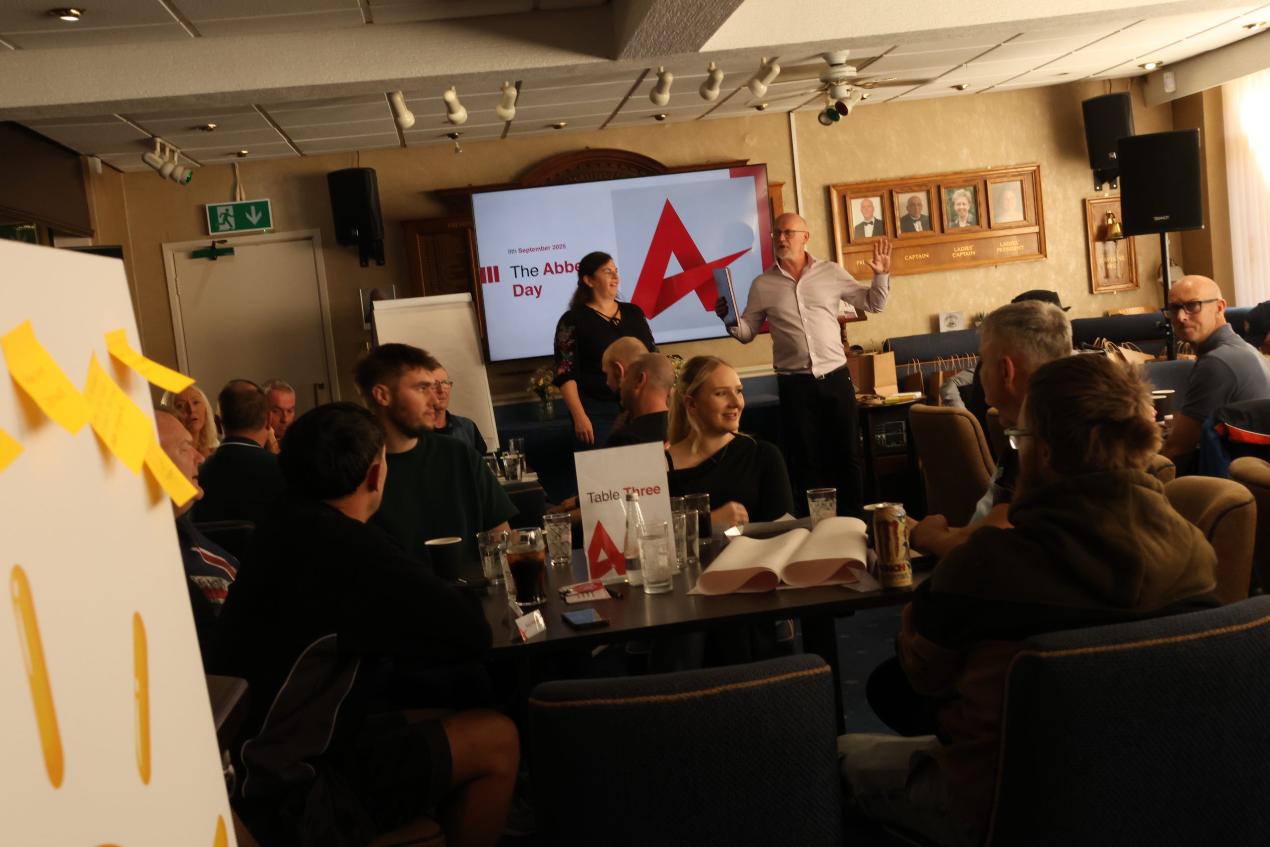 A collection of square tables with people sitting at them at smiling with a large TV behind them with 'The Abbey Day 2025' emblazoned onto it