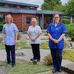 bury hospice staff members are dressed in blue nursing uniform and standing in a small garden surrounded by plants with a large red bricked building in the background