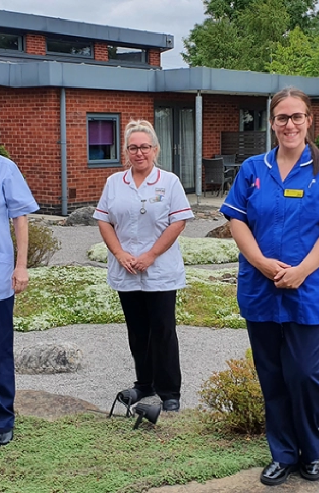 bury hospice staff members are dressed in blue nursing uniform and standing in a small garden surrounded by plants with a large red bricked building in the background