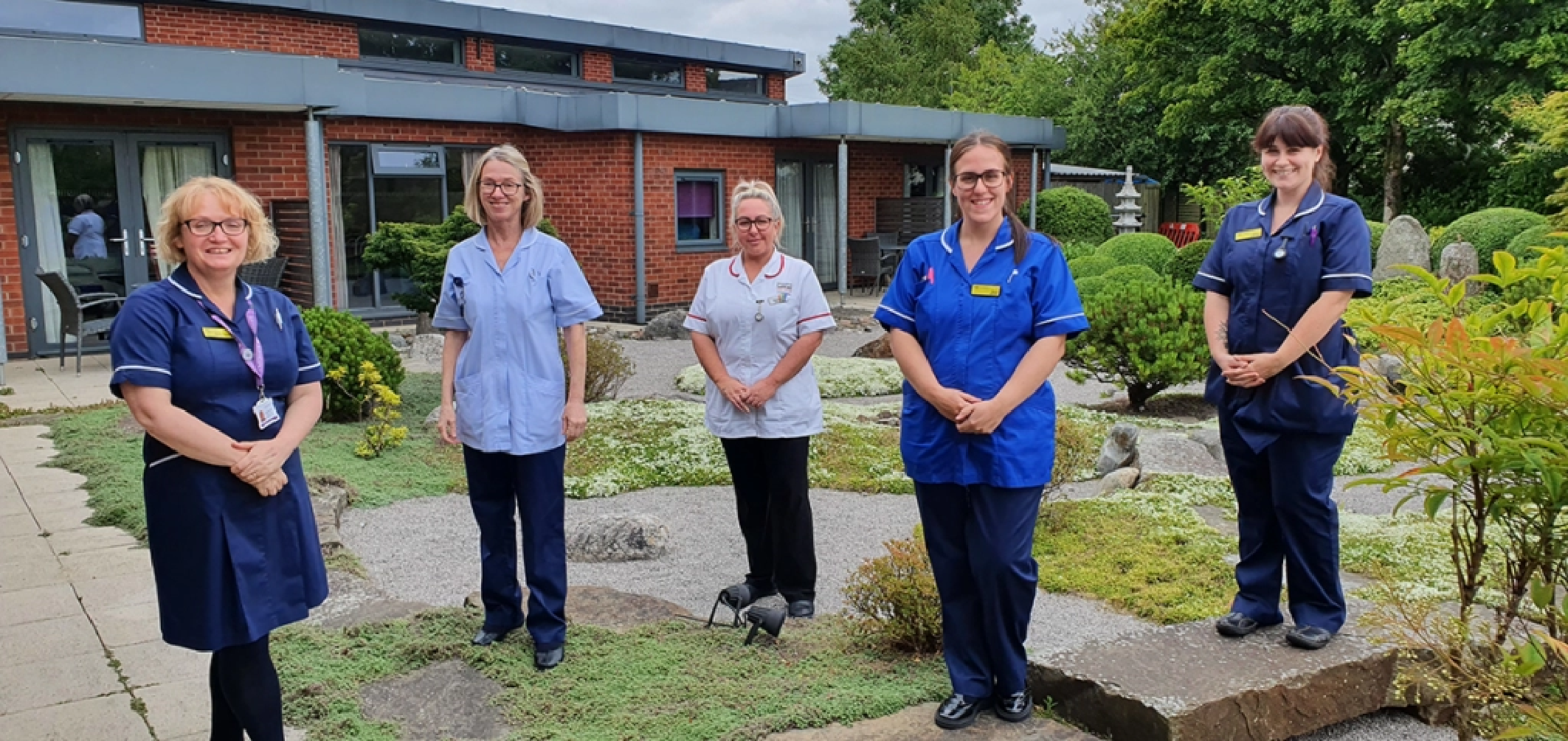 bury hospice staff members are dressed in blue nursing uniform and standing in a small garden surrounded by plants with a large red bricked building in the background