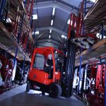 A man is driving the abbey forktruck in a warehouse surrounded by large racking