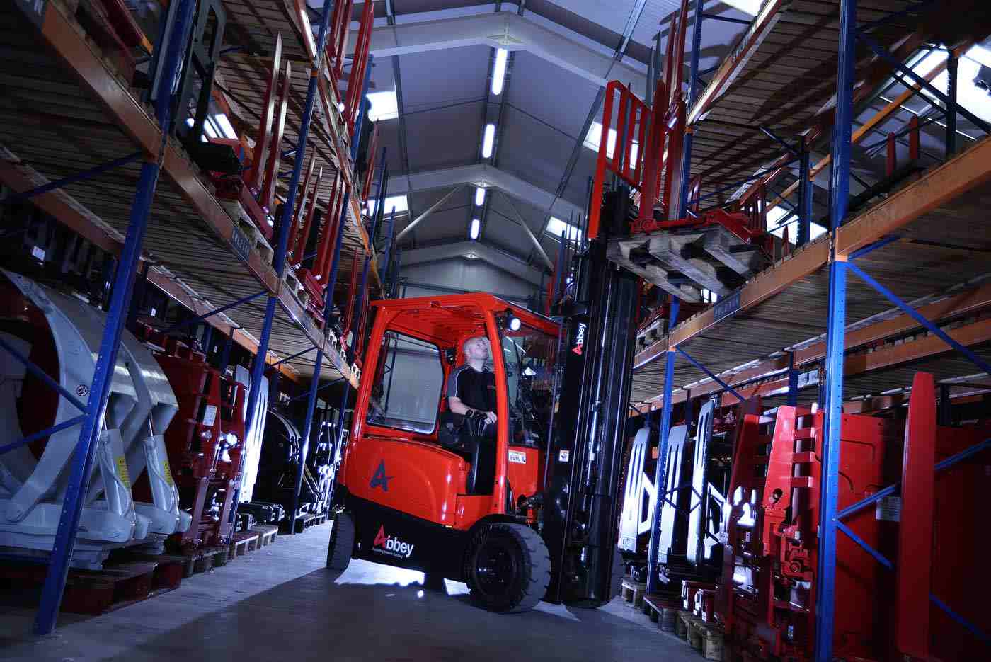 A man is driving the abbey forktruck in a warehouse surrounded by large racking