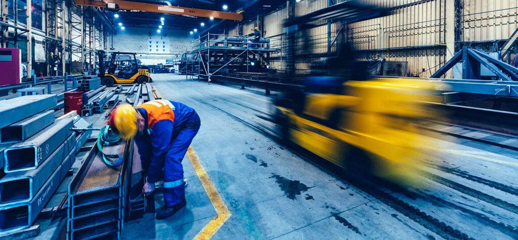 a warehouse worker in a orange hi-vis is working a steel plate while a blurred yellow forktruck drives behind him