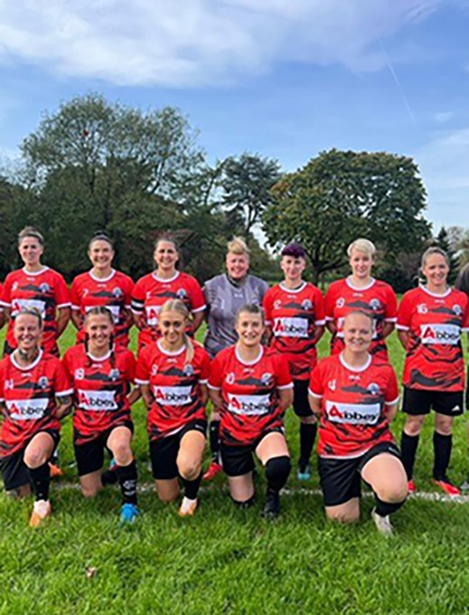 the entire JPS ladies FC squad are posing in on a football pitch wearing the red JPS jersey
