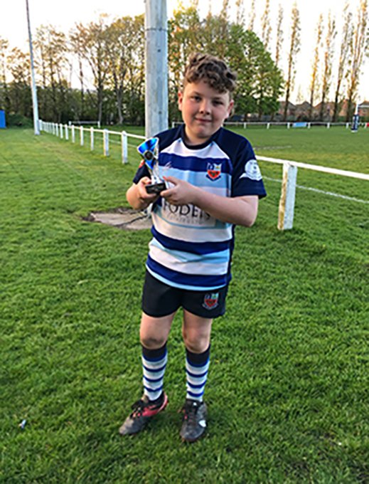 a junior Winnington Park rugby player is wearing the navy and blue jersey holding a trophy in front of a rugby pitch