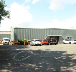 A rectangle warehouse with a grey, cladded roof and a few cars parked outside