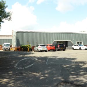 A rectangle warehouse with a grey, cladded roof and a few cars parked outside