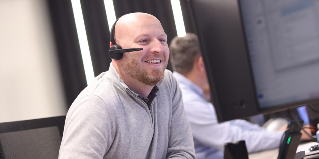 mark anderton is wearing a headset, wearing a grey jumper, and smiling at a computer screen